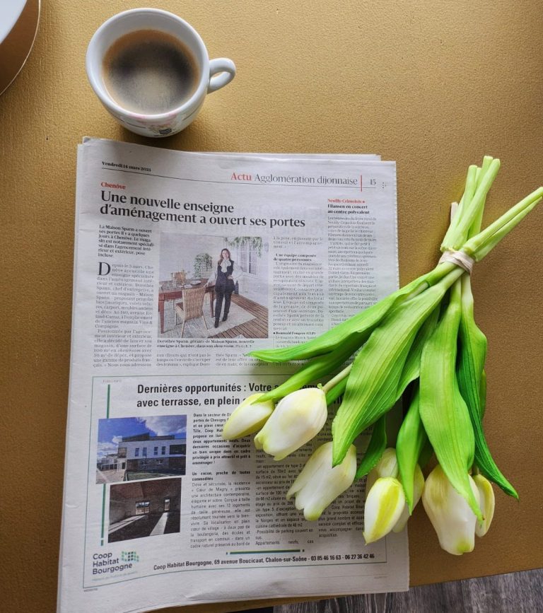 Article du Bien Public - Maison Spann Un journal ouvert, une tasse de café et un bouquet de tulipes blanches sur une table.