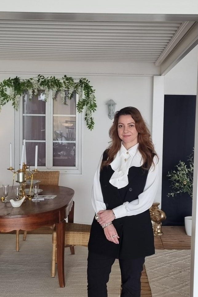 Maison Spann - agencement intérieur et extérieur à Dijon Femme debout dans un magasin d'exposition, sous une pergola blanche Solisystème, avec une table en bois et des plantes décoratives.